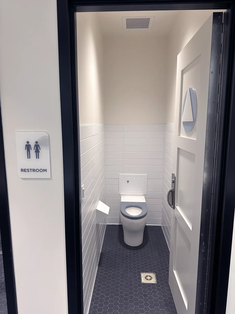 Restroom at the Be Well OC campus with a BestCare solid surface toilet, gray integral seat and flush valve access panel, seen through the open door